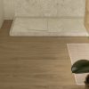 Suelo Dekton Kedar en baño con plato de ducha blanco, alfombra y planta, en tono roble cálido.