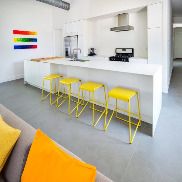 Isla blanca Neolith Arctic White con taburetes amarillos, arte arcoíris y electrodomésticos en cocina abierta.