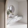 Vanitorio flotante Neolith Azure blanca veteada con espejo redondo, grifo dorado y planta en baño luminoso.