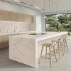 Cocina moderna con isla Ceratop Macadamia Cream crema veteado, gabinetes blancos, luces lineales y vista ventana en ambiente luminoso.